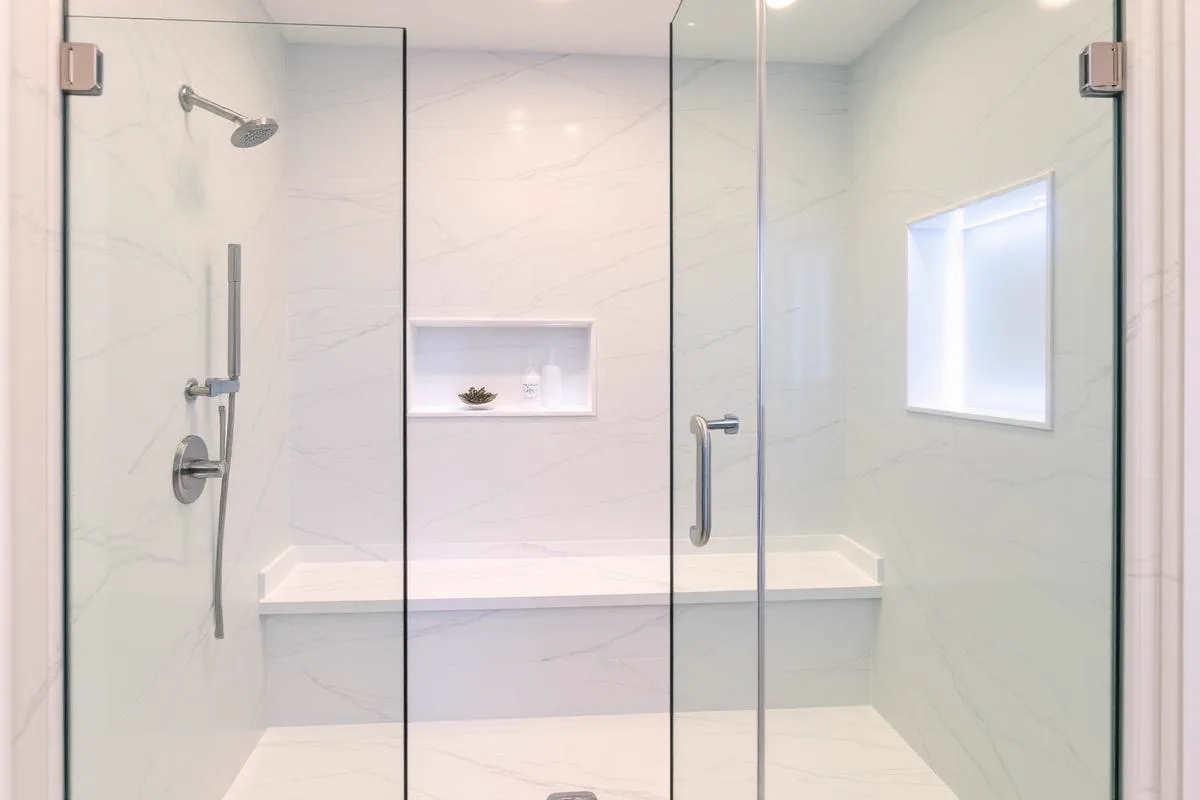 The inside of a white marble shower, with frameless glass panels and a door with a chrome handle, a chrome rain head and handset, a full-width bench, and two recessed niches, one of them lit